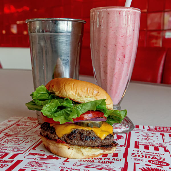A cheeseburger, strawberry milkshake, and metal shake cup on a diner counter with red accents.