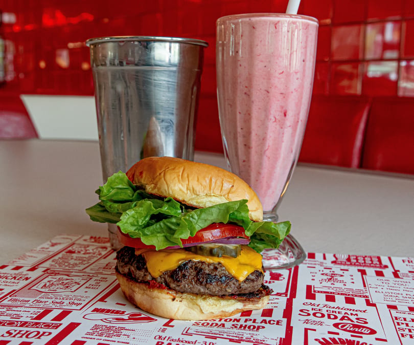 A cheeseburger, strawberry milkshake, and metal shake cup on a diner counter with red accents.